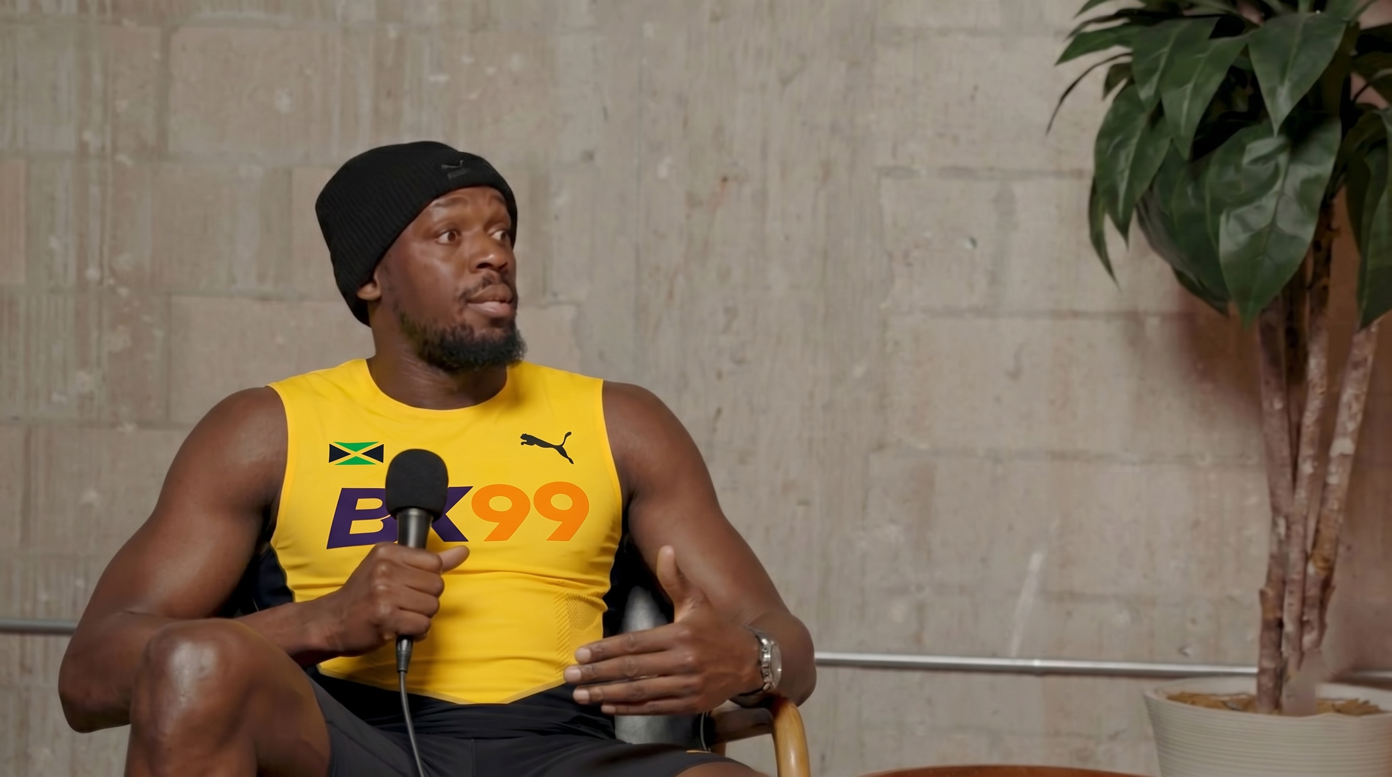 Usain Bolt sitting in a leather chair wearing a yellow BX99 official jersey, talking to the camera and holding a BX99 script