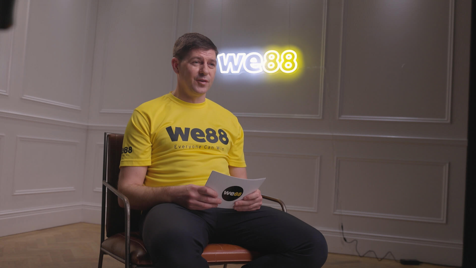 Usain Bolt sitting in a leather chair wearing a yellow BX99 official jersey, talking to the camera and holding a BX99 script